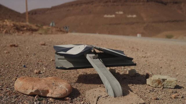 Mangled metal road sign bent and crushed on roadside ground beside rocks with rocky desert hillside in southern Morocco