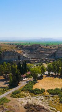Vertical video. Balat, Turkey. The ancient Theatre of Miletus, a vast semicircular stone amphitheater with tiered seating carved into a hillside, aerial drone view under clear summer sky. Aerial View