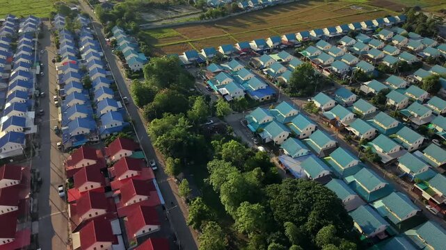 A smooth, high-angle aerial drone shot revealing a master-planned housing development with rows of red and blue roofs transitioning into lush, green rice paddies and rural landscapes at dawn. 
