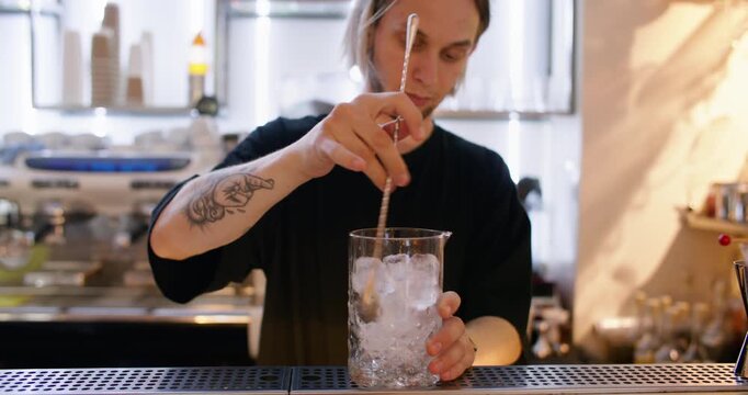 Cocktail preparation. Cold drink. Bartender stirring ice cubes in glass with long bar spoon making chilling ingredient on pub counter.