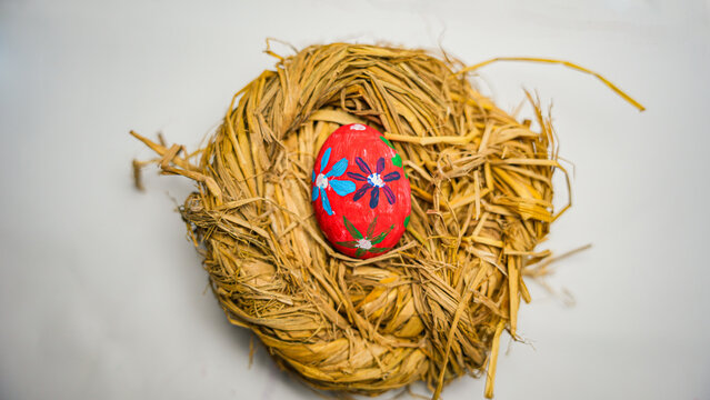 A single, red Easter egg with a detailed floral pattern is centrally placed in a handmade straw nest, casting a soft shadow on the white background.
