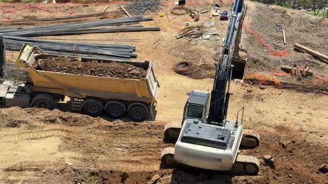 Crawler excavator loading soil and stones into the tipper of a white dump truck at an open construction site. Heavy machinery logistics field operation.