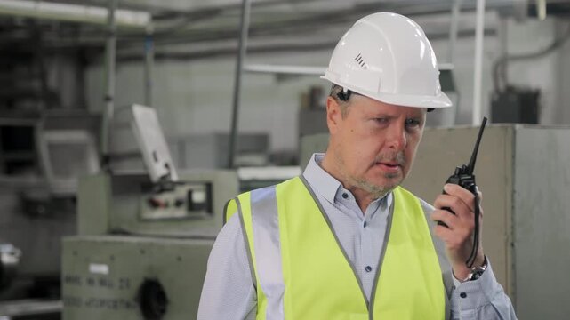 Factory engineer in safety helmet and reflective vest communicating by walkie talkie during industrial production process.