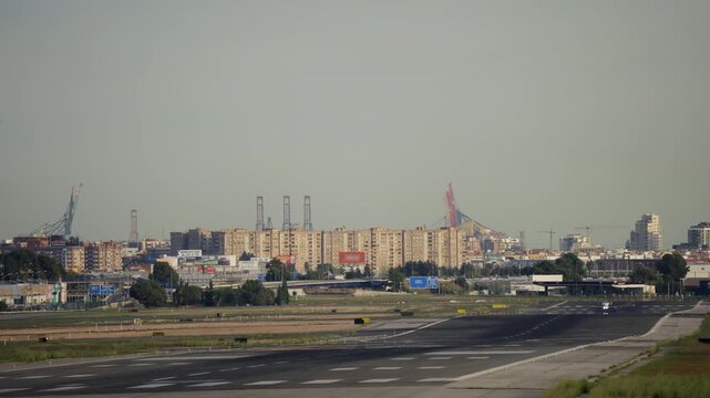 Wide airport runway view in morning haze with city skyline and cargo port cranes in distance, small aircraft is taking off