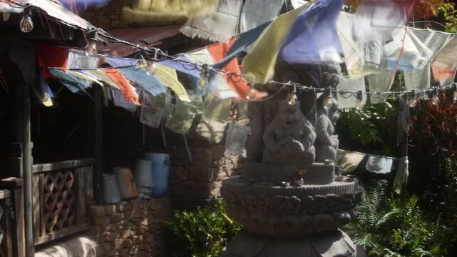 Prayer flags hanging above a stone statue in a sunlit temple garden. Rustic wooden railings, brick walls, string lights, and greenery frame the cultural courtyard scene