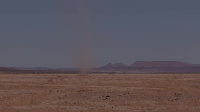 Thin dust devil whirlwind rising from a flat dry desert plain against hazy mountains and grey sky Morocco