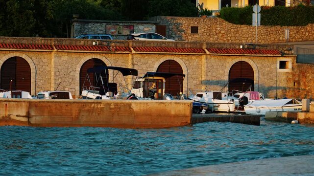 Traditional small boats docked beside stone waterfront architecture on Krk island in Croatia during warm sunset light along the Adriatic coastline.