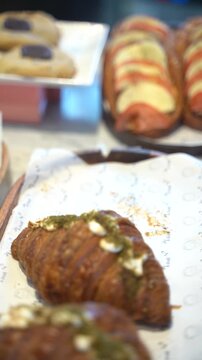 Food service action. A gloved hand lifts a freshly baked, golden croissant decorated with savory toppings, with caprese sandwiches blurred in the background.