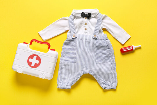 Toy first aid kit with kid's clothes and thermometer on yellow background. Top view