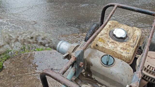 A dirty gas-powered motor pump actively pumping and discharging a strong stream of muddy water onto the concrete ground during drainage or emergency utility works