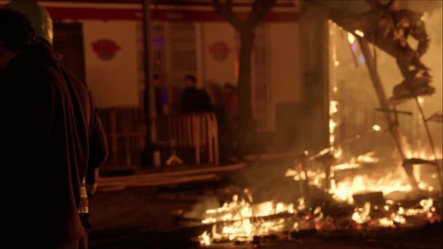 Firefighter in reflective jacket and helmet stands back to camera watching burning falla during Las Fallas crema in Valencia, Spain