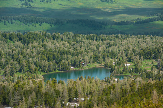 Scenic aerial top view to wood lake against woody hills. Colorful view with cabin on shore of forest lake. Sunlight and shadows of clouds on hilly vastness. Coniferous trees reflection in alpine lake.