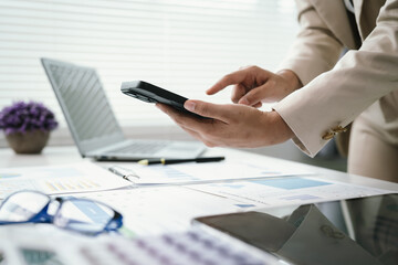 Fototapeta premium Close-up of a business professional using a smartphone while reviewing financial documents at a modern office desk, highlighting mobile communication, productivity, and digital workflow.
