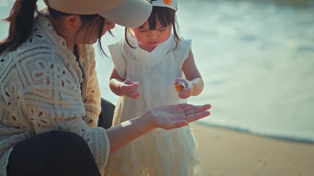 Caring Asian mother and cute little daughter collecting seashells on sandy beach during summer vacation, happy family playing together near sea, child development, outdoor learning and travel concept