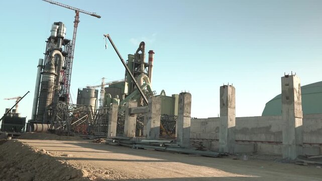 Wide shot of an industrial cement production site under development, featuring large concrete pillars, massive steel scaffolding, and heavy-duty cranes in operation