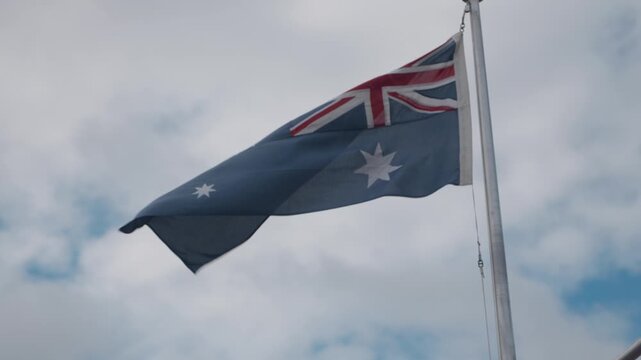 An Australian flag from the back flying high on a flag pole, blowing in the wind with the blue sky and white clouds in the background.