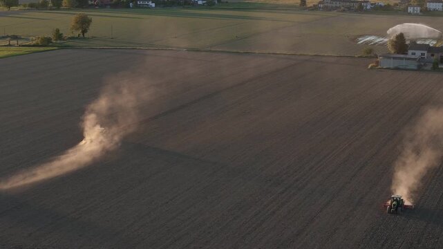 Agricultural tractors perform harrowing and seedbed preparation across drought affected farmland in Province of Piacenza Emilia Romagna Italy creating drifting dust plumes, slow oblique drone slide