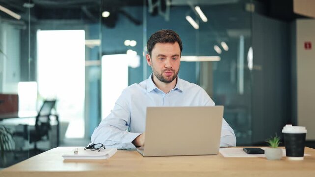 Male web developer or freelancer talking on webcam video conference. Professional remote worker speaking and gesturing at laptop screen during online business meeting at office desk.