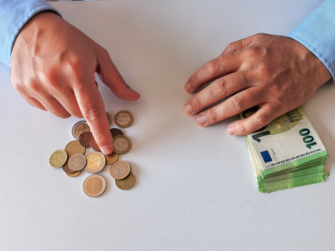 A man in a blue shirt counting cash (Euro currency). A stack of 100 Euro banknotes and scattered Euro cent coins are on a white table. 