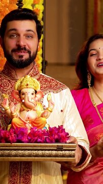 Indian Couple Welcoming Ganesh Idol with Joy, Young husband wife in Traditional Attire Holding Ganpati Bappa Idol, Puja Thali While Smiling and Chanting Bappa Morya During Ganesh Festival Celebration