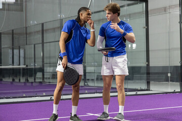 Obraz premium Diverse male partners talking and gesturing on purple court in blue shirts holding paddles