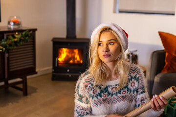 Fototapeta premium Adult woman sitting in living room near wood stove holding wrapping paper roll, wearing Santa hat