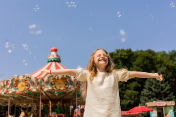 happy little girl is blowing bubbles in front of a carousel at an amusement park. © Tina kids photo