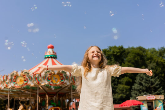 happy little girl is blowing bubbles in front of a carousel at an amusement park.