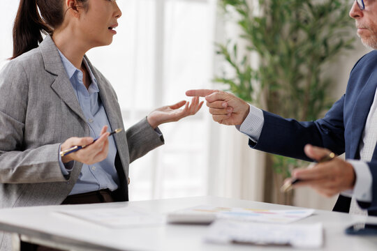 Business colleagues in heated office discussion: woman gestures with pen while older man points accusingly, capturing tension, miscommunication and workplace conflict