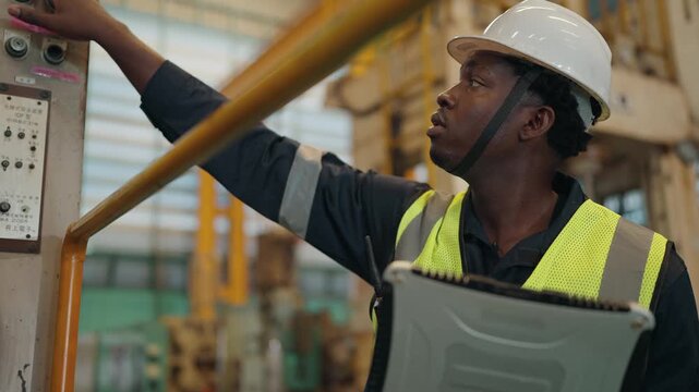 Factory worker at machine: A male factory worker in a white hard hat and safety vest operates a complex control panel, his hand adjusting a dial with focused intent.