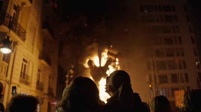 Silhouetted crowd on city street watching massive falla sculpture consumed by towering flame during traditional Fallas fire festival in Valencia, Spain