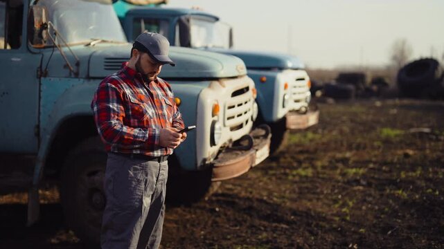 Farmer and agricultural machinery in farmland, medium portrait in harvest season . Bearded man in checked shirt using modern smartphone, testing new mobile application for farmers and agronomist