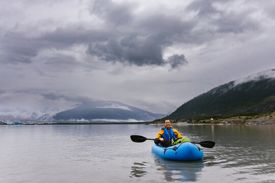 Man smiling in blow up kayak on glacial lake with cloudy sky