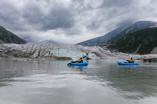 Two people paddle blow up kayaks on glacial lake with cloudy sky