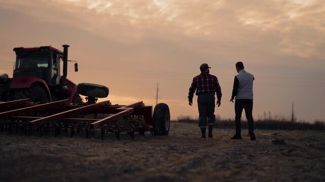 Farmer and agronomist walking in agricultural fields in sunset, back view. Agribusiness and agronomy, professionals discussing modern machinery for farming and soil processing in ecological region