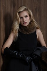 Naklejka premium Blonde woman posing in dark grey winter coat with fur collar, holding black leather gloves near face in studio setting. fashion, winter, portrait, elegance, style