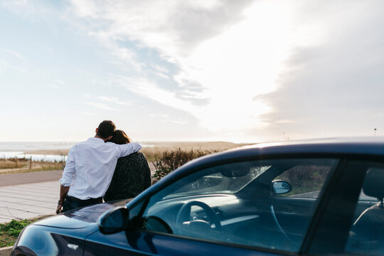 Spain, Cubelles, back view of a young couple sitting on the hood of the car watching the sea at sunset