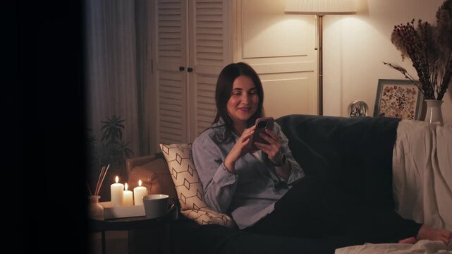 Happy woman using smartphone at home in cozy living room. Smiling young woman subscribing to new social media, shopping online, ordering products online using apps, watching series, chatting with