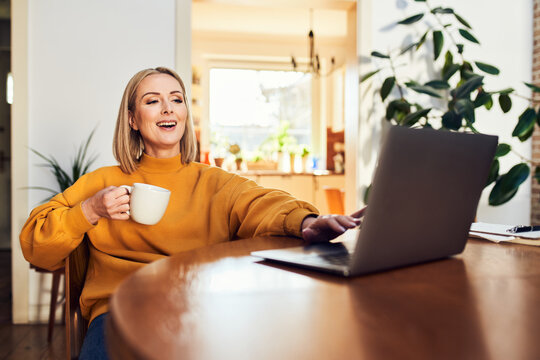 Woman using laptop and drinking coffee at home dining table