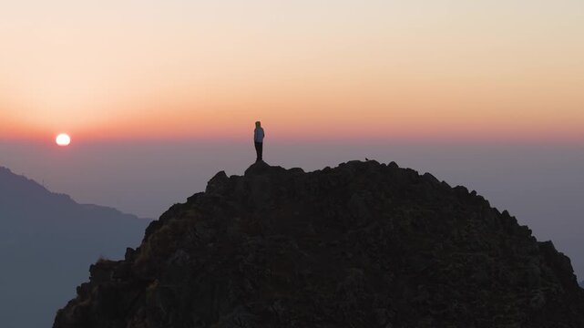 4K drone shot of a young man standing on a rugged Himalayan cliff during colorful morning sunrise.