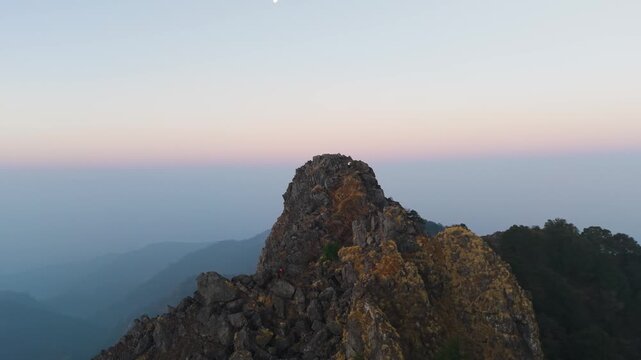 4K aerial view of a boy hiking up a rocky cliff with colorful sunrise skies in the Himalayas, India.