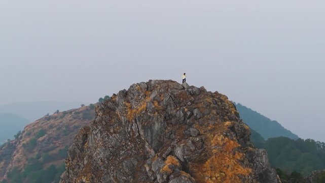 4K aerial 360 drone shot of a boy standing on a rocky mountain peak during sunrise in Uttarakhand, India.