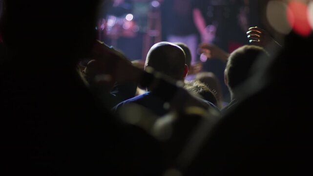 Concert fan claps hands held overhead in crowd, wristband visible, blurred pink and purple bokeh lights fill background, performer dimly visible on stage