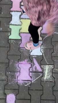Child playing hopscotch on pastel chalk tiles, preschooler in pink jacket and cartoon slippers hops between colorful squares on urban brick pavement,