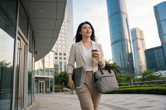 Businesswoman walking outdoors with coffee cup and laptop bag in modern city environment