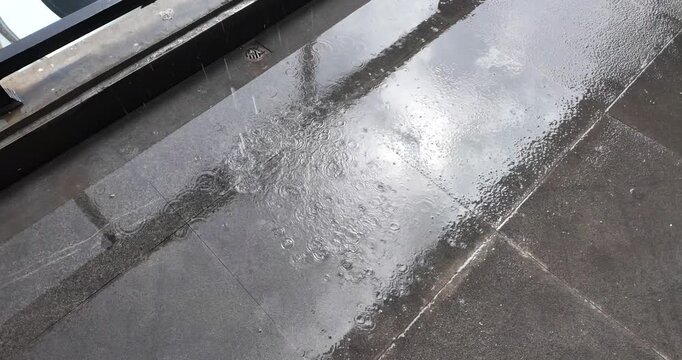 Slow motion of raindrops falling onto a wet, dark tiled surface, creating concentric ripples while reflecting the light and sky above.