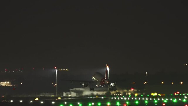 Commercial airliner on final approach descending over colorful green and red runway and taxiway light at night, terminal building and floodlight visible in dark background
