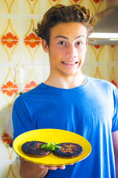 A teenage boy in a blue t-shirt holds a yellow plate with two burnt pancakes, making an awkward nervous grin in a retro tiled kitchen, conveying surprise and a cooking mishap
