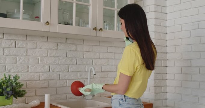 Young girl talking by the phone advising how to unclog a sink drain. Girl trying to clean sink using plunger standing in the kitchen at home. Housekeeping and household work concept. 4k video.
