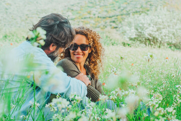 Obraz premium A smiling couple sits together in a wildflower meadow, sharing a warm moment surrounded by green grass and white blossoms, evoking love, relaxation and outdoor springtime joy.